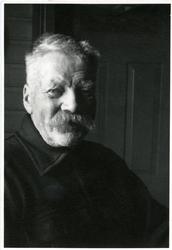 Portrett av en eldre mann sittende foran en vegg og dør. Man