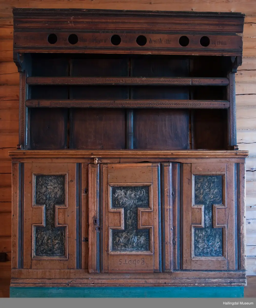 Rester etter rød og blå maling.  Gesims, skråner ned bakover. Profilert. 6 sirkulære hull i arkitraven.  Mellom disse er malt med sorte bokstaver.  Gavlene har utskårne "hylleknekter".  Bakveggen består av fire vertikale planker.  Skjøtene er dekket med profilerte lister.  Bare to biter i nederste del til venstre er fremdeles på plass.  Nederste del er lukket skap.  Tredelt, midterste del er skapdør.  Platen har profillist.  Feltene har speilfyllinger.  Blåmalte.  Klosset marmorering.  Speilfyllingenes form er som en liggen H.  Har profillist.  Skapet er satt på en ny grønmalt plate.  På døren er malt hvite bokstaver.  Nederst på døren er skåret inn bokstaver, opp-ned.  To nøkkelhull m. jernbeslag på venstre side av døren.  Over døren til venstre, en dreibar trekloss, holder igjen.  Innvendig har døren merke etter stort jernbeslag på samme side som nøkkelhullene.  To lister skåret i treverket minner om lange jernbeslag,-smalner mot nøkkelhullene.  Akantusranker i rødt, blått og hvitt, meget avslitt.  En hylle i skapet. Mangler bakvegg.  Karmen har på venstre side spor etter to jernhengsler.  Øverst på høyre kortside, mangler gesimsen en bit, og holdes til sideveggen med en jernkrampe.  Jernkrampe også på venstre side.  Skapplaten mangler list på høyre side.  Venstre side, fra gesimsen og ned har spor etter å ha stått inntil en vegg eller et annet møbel.