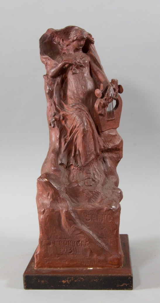 Kvinna, Sappho, stående håller en trälyra med metallsträngar i ena handen. I andra handen, tryckt mot bröstet, blommor. Lutad mot klippformation.