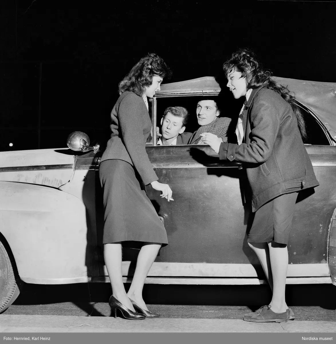 Gatumiljö med bilburen ungdom.Två unga kvinnor, den ena med cigarett i handen, hänger vid bil och samtalar med två unga män som tittar ut genom den nedvevade rutan. 