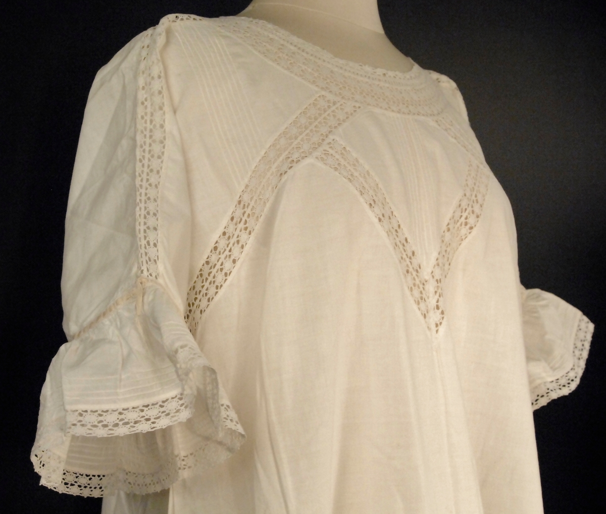 Dekorert med biser, blonder og smale gule silkebånd. Kort erme med kappe. Utskrådd kjole med kiler i sidene nederst.