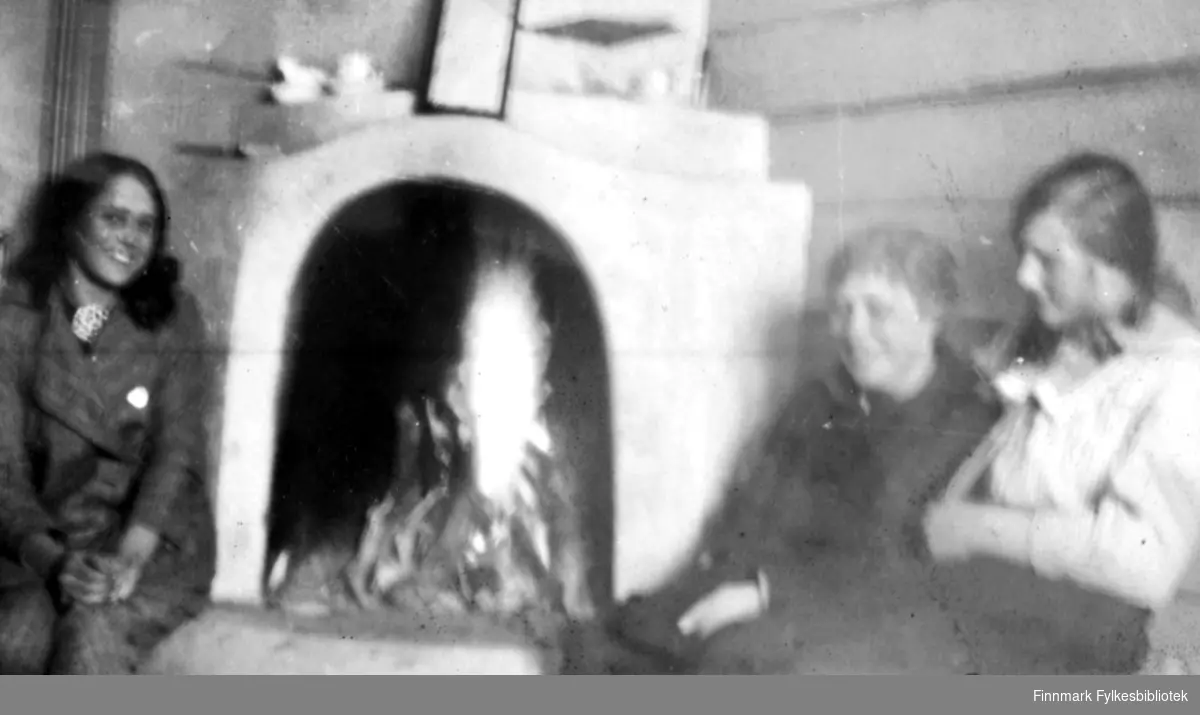 Grønlien, mai 1917. Tre kvinner sitter rundt en peis ved bålet. Til venstre en yngre kvinne i sportslig drakt med en stor brosje eller sølje i halsen. Til høyre en litt eldre dame nærmest bålet og en ung pike sitter ved siden av. Ref. også FBib.01010-116 som viser at dette er en hytte. Bildet er bevegelsesuskarpt, men vi kan se gjenstander som står plassert oppå peisen.