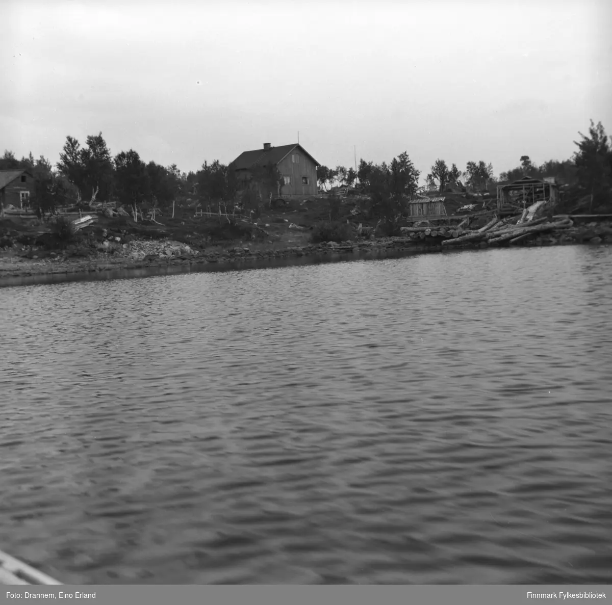 Bilde tatt fra innsjøen mot et hus og noen hytter langs bredden.