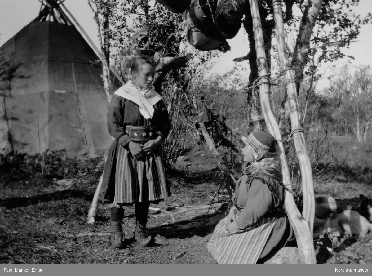 Inga Prost i silverknappsbälte och sidensjalett frågar sin mamma om lov att fotografera sig. I bakgrunden en kåta, i ett träd hänger korgar. Bilden tagen i Viikusjärvi, Vittangi sameby, Kiruna.