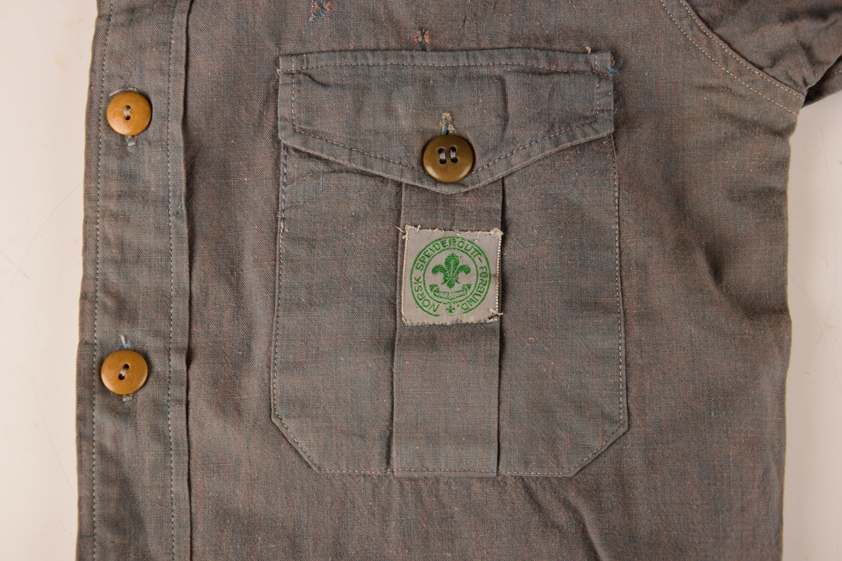 Speiderskjorte i grønn tekstil.

6 brune plastknapper, støpt med fransk lilje som motiv. To brystlommer. Knapp ved hvert erme. Påsydde merker.