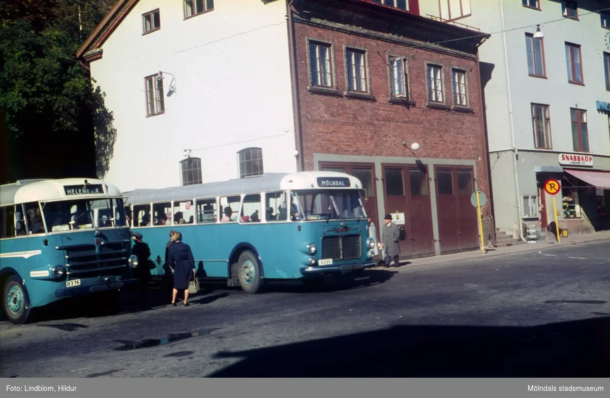 Två blå bussar står på en busshållplats vid Gamla Torget i Mölndal, 1950-60-tal. I mitten ses huset Kvarnbygatan 41, tidigare brandstation och garage för polisbilar, numera Mölndals målarskola. Till höger ses del av huset Kvarnbygatan 43, tidigare Mölndals stadshus, sedermera musikskola.

För mer information om bilden se under tilläggsinformation.