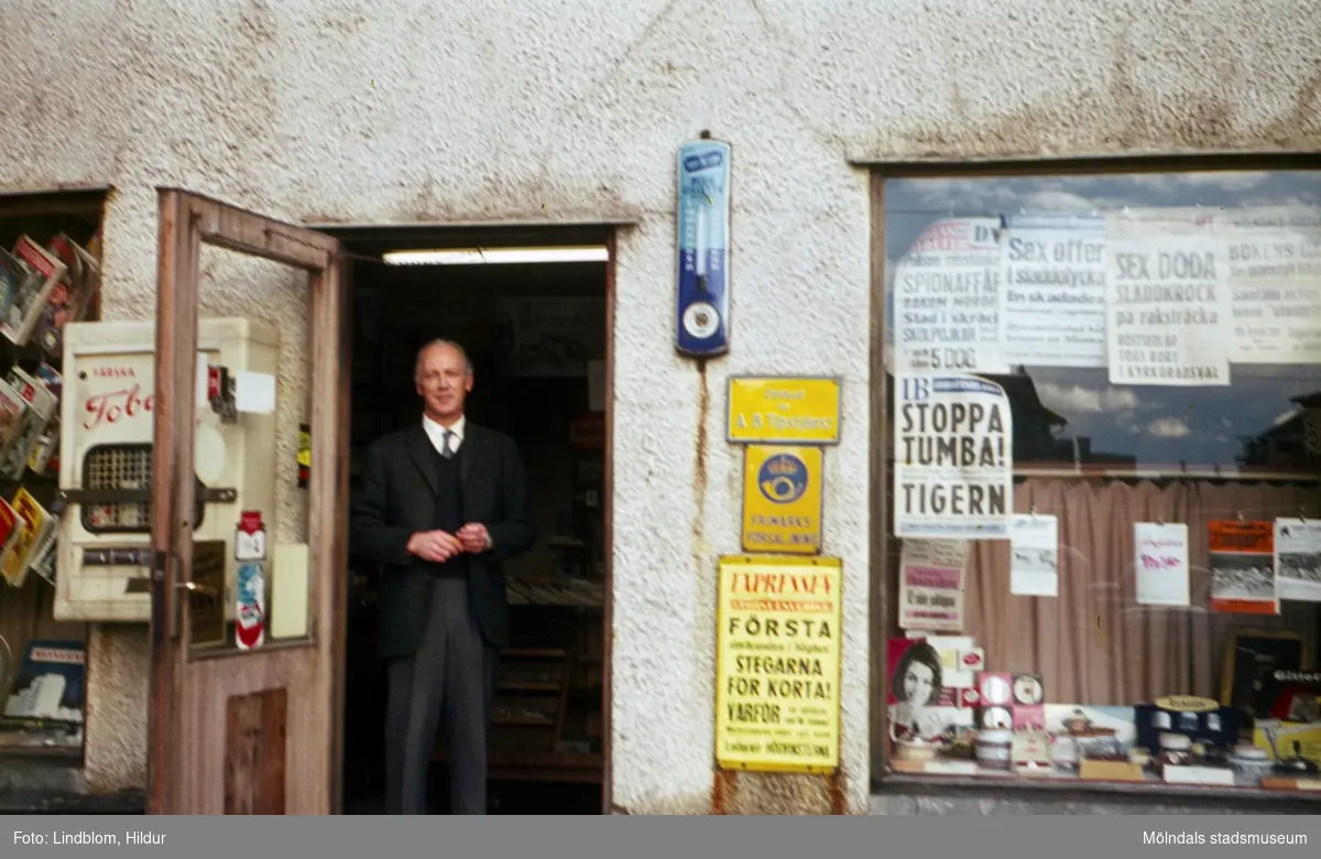 En man ståendes i dörren till tobakshandeln med adress Kvarnbygatan 43 vid Gamla Torget i Mölndal, 1960-tal.

För mer information om bilden se under tilläggsinformation.