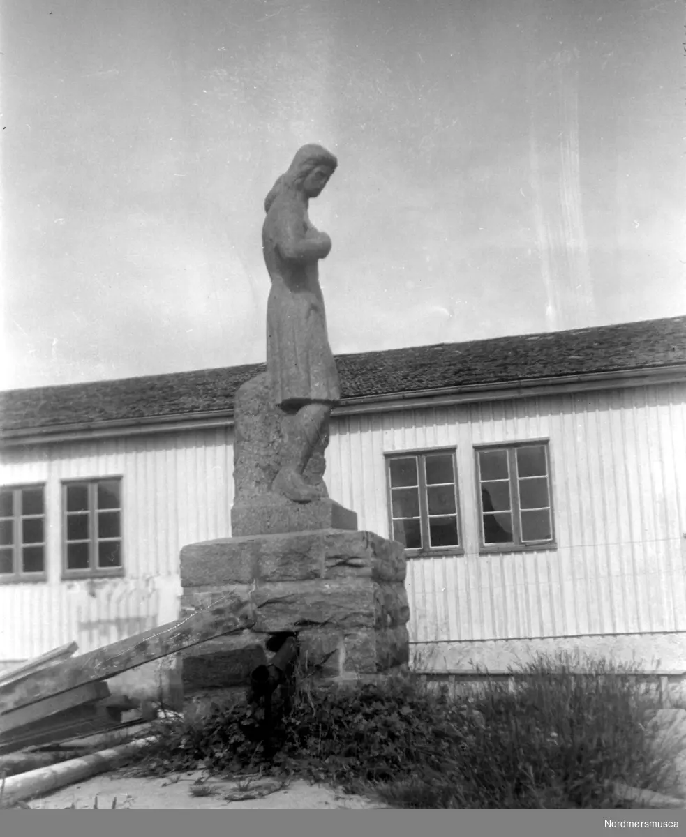 Nordre fløy av Soldatheimen med statuen i forgrunnen, i Lars Guttorms gate - nær Allanengen Skole. Statuen er utført av en tysk soldat ved navn Rainer, og forestiller en ung kvinne. Rainer, som var steinhugger og treskjærer i det sivile, laget statuen i en brakke i Vågen. Et vakkert kunstverk.
Etter 1945 ble bygningen solgt til Kristiansund Arbeiderparti, som til per dags dato (august 1955) har brukt lokalet som samlingssted, møtelokale, samt til utleie for byens forskjellige foreninger. Det har også blitt brukt til offentlig valglokale (den store salen). I disse dager (per august 1955) holder de på å rive Soldatheimen, for å gi plass til byggetomter. (Metodistkirken). Kilde (blant andre): Tidens Krav, Fredag 14. september 2007, side 20

";Stein Bach påsto at det var to slike statuer, den største stod utenfor "Soldatheimen" og en mindre sto inne i vestibylen. Han kunne fortelle at den største som ble knust og fraktet sammen med mer stein derifra til plassen uten for huset til far til Stein (Mellomveien 7 på Steinberget), da brukt som fyllmasse (noen påstår at dette ikke er riktig og det var bare en statue). Den gjenværende statuen står i hagen til tidligere byingeniør Røkkum. Den står der fremdeles. Det er i bakhagen til det øverste huset i Omveien. Du kan se den fra Nord-østliste delen av Kringsjå. Når du går fra Omveien opp til Kringsjå så ser du den til høyre."; Ut i fra dette så er den kanskje lagt ned under parkeringsplassen i Mellomveien, men det er ikke sikkert den er hel.



Bildet er fra Nordmøre Museums fotosamlinger.
