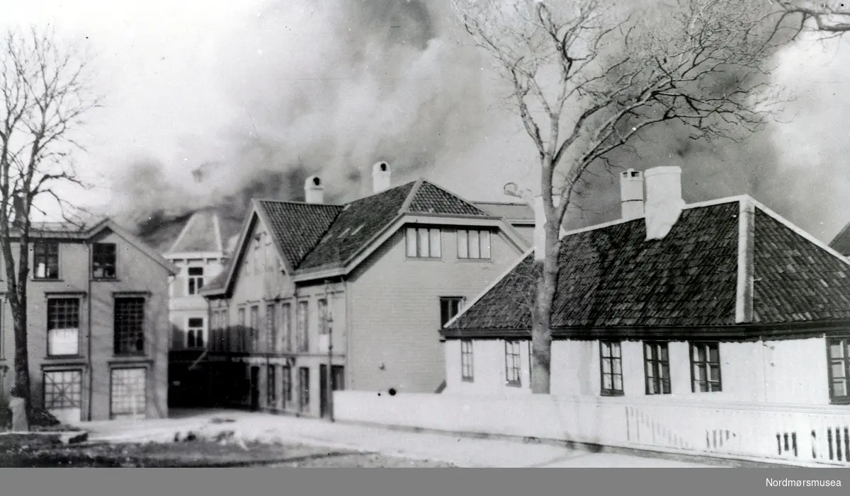 Foto fra Storgata på Kirkelandet i Kristiansund, hvor vi ser et av de nære husene til Knudtzongården (til høyre) i brann etter at bombingen av byen har startet i april 1940. Fra Nordmøre museums fotosamlinger.
