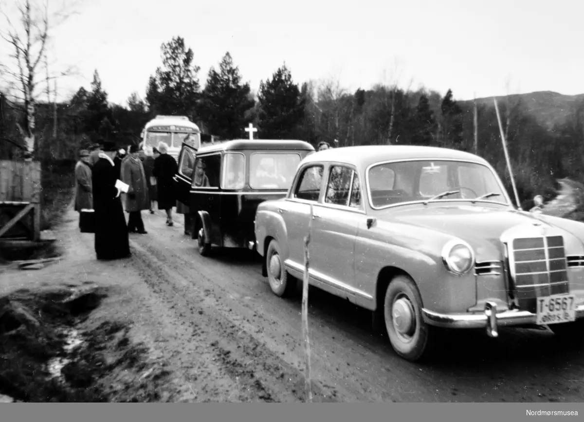 Foto fra et gravfølge. I front ser vi en bil med registreringsskiltet T-6567. Like bak er bårebilen og gravfølget med prest. Trolig er følget foran en ukjent kirke på Nordmøre, i Møre og Romsdal. Dateringen: Bilen, en drosjeregistrert Mercedes-Benz, er av en modell som ikke kom før i 1955. Bårebilen er ikke det, men en tilhenger - en ikke uvanlig løsning i landdistriktene på den tid. Bakerst en buss. Info: Ivar Stav.  Ukjent fotograf. Fra Nordmøre Museums fotosamlinger.
