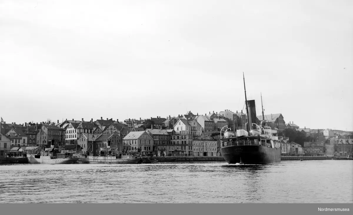 En rutebåt liggende ute i havnebasenget, på Kirkelandet i Kristiansund. Vi ser også to ferger ?? liggende ved kai, ved Piren. Nordmøre Museums fotosamling