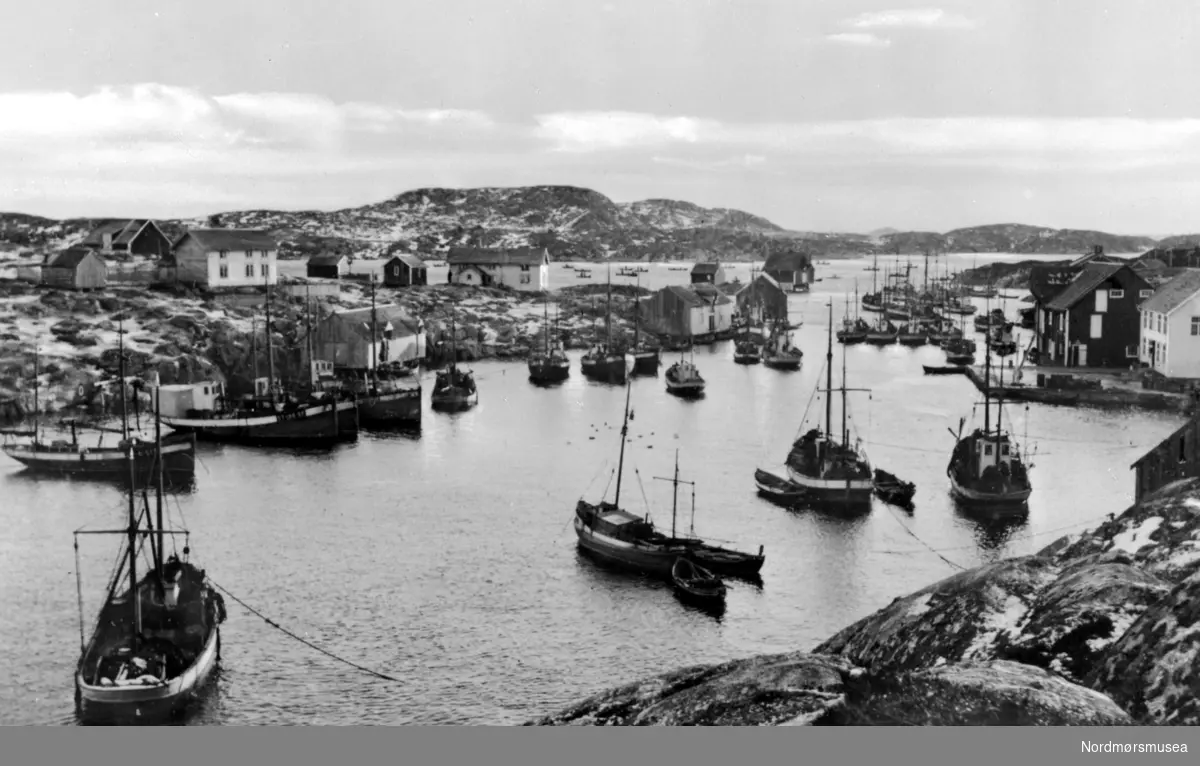 Foto fra båthavnen i Bogøyvær i Frøya kommune, Sør-Trøndelag fylke. Her ser vi flere fiskebåter liggende fortøyd inne i den lune havnen, med deler av bebyggelsen liggende i rundt. Fotograf er trolig Georg Sverdrup, og datering er sannsynligvis mellom 1930 til 1930. Fra Nordmøre Museums fotosamlinger.