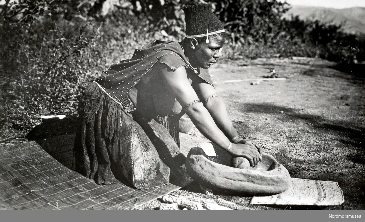 En afrikansk kvinne kverner mais med en skubbekvern. Fotograf, sted og datering er ukjent. Fra Nordmøre Museums fotosamlinger. EFR2015