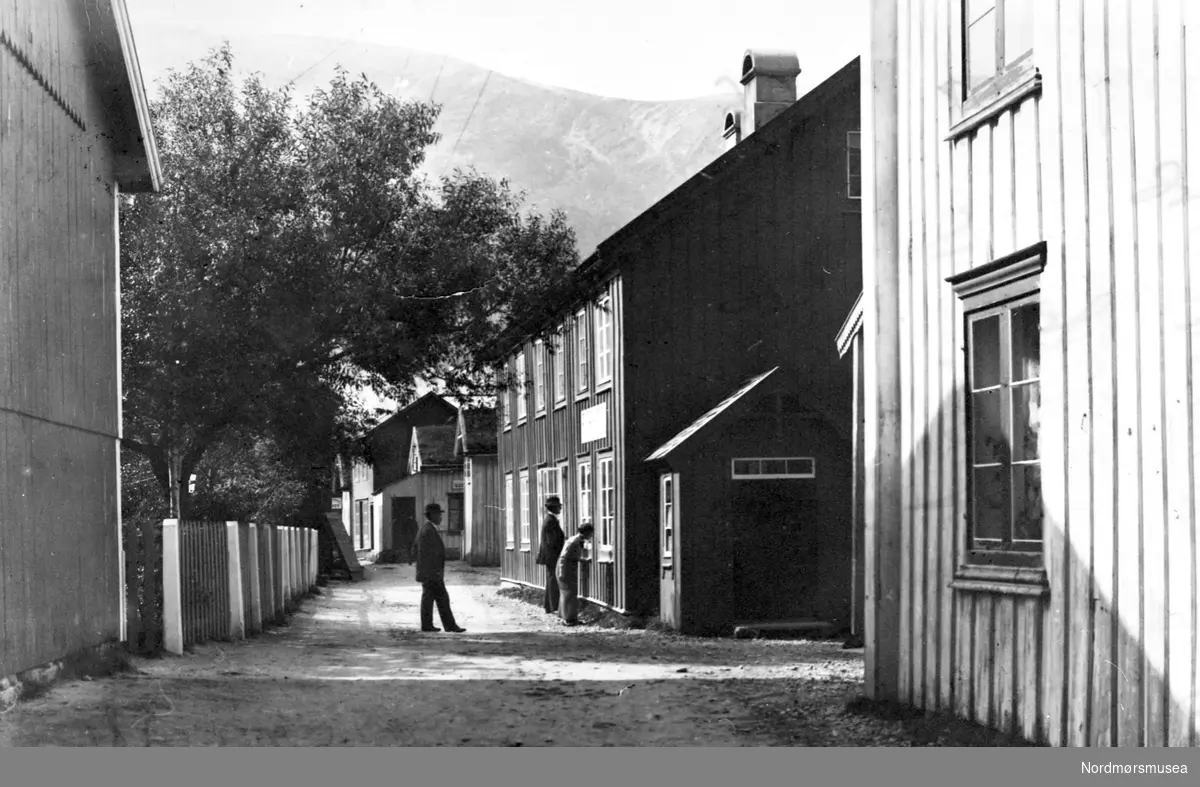 3 menn kikker inn i et butikkvindu et sted med høye fjell bak. Sunndalsøra? Gate, hus,bygning. ca 1890. Fra Nordmøre Museum sin fotosamling.
