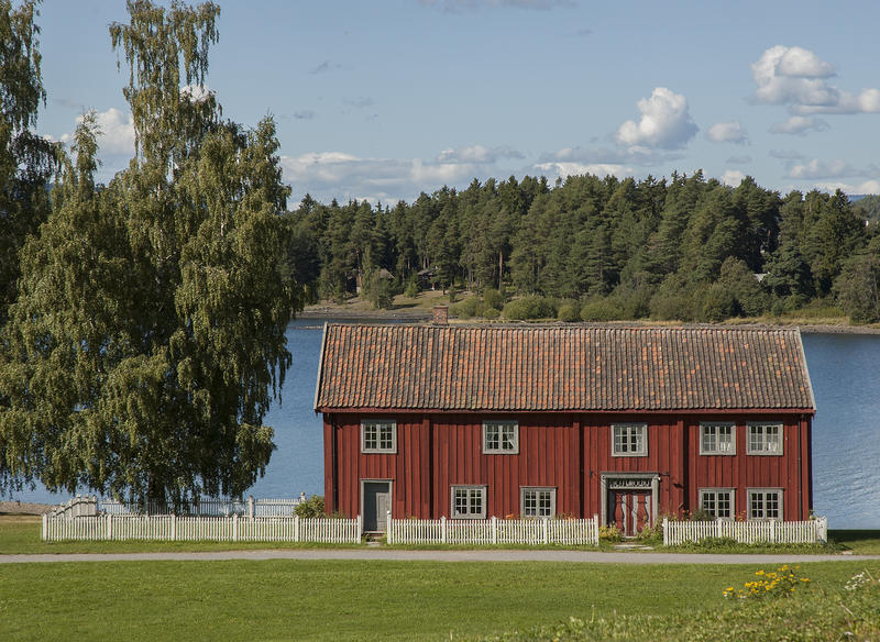 Stor, rød bygninge i to etasjer ligger bak et hvitt stakittgjerde ned mot vannet. På venstre side to bjørker. (Foto/Photo)