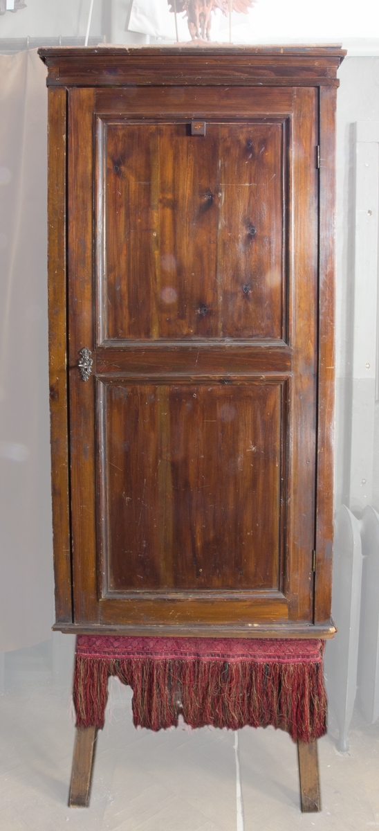 Skåp, verktygsskåp, av betsad furu, stående rektangulärt med dörr och fyra hyllplan. Dörren har utvändigt profilerade lister. Krönlist. Nyckel och profilerad nyckelskylt av metall. Skåpet står på ett fyrbent underrede av trä.