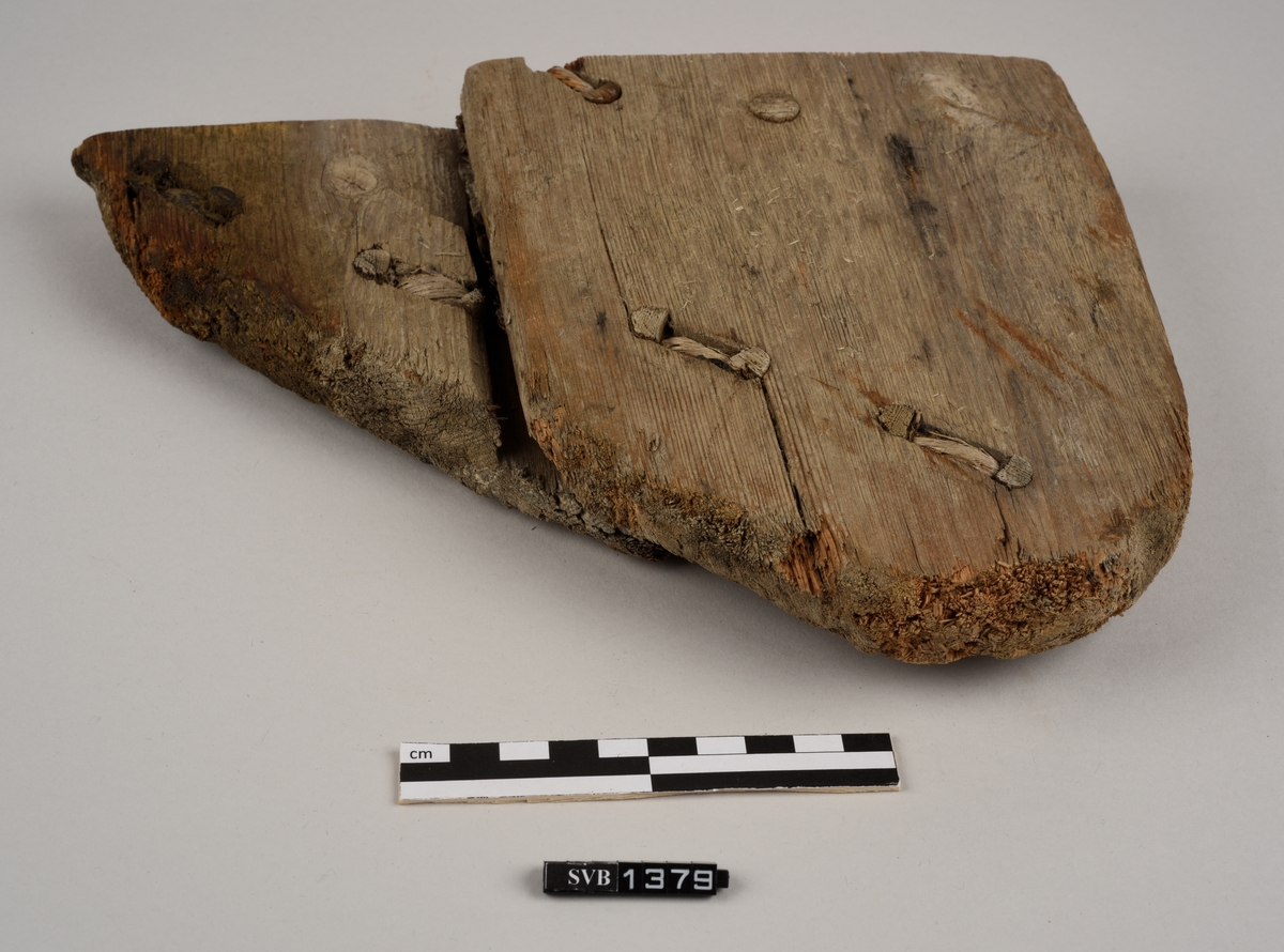 Del av et båtbord. Delen ble saget av. Det er hull til vidje og plugger. Delvis er det rester av både vidje og plugger. Treet fremstår ubehandlet. Det er et materiale som ligner torv/mose mellom bordene.