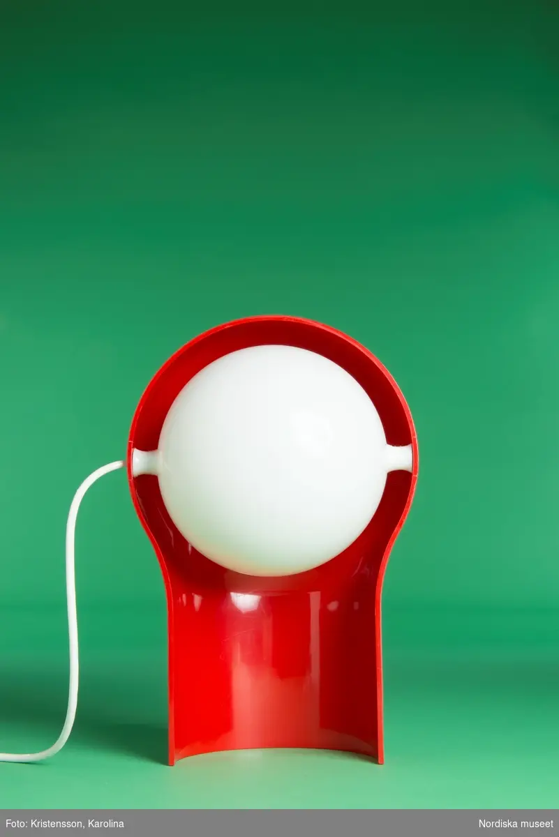 Bordslampa av ABS-plast, orange skärm med halvsfärisk, ställbar reflektor i vitt. Inuti skärmen lampsockel för kronljusglödlampor. Vit plastsladd från lampans mittparti med strömbrytare och stickkontakt.
/Maria Maxén