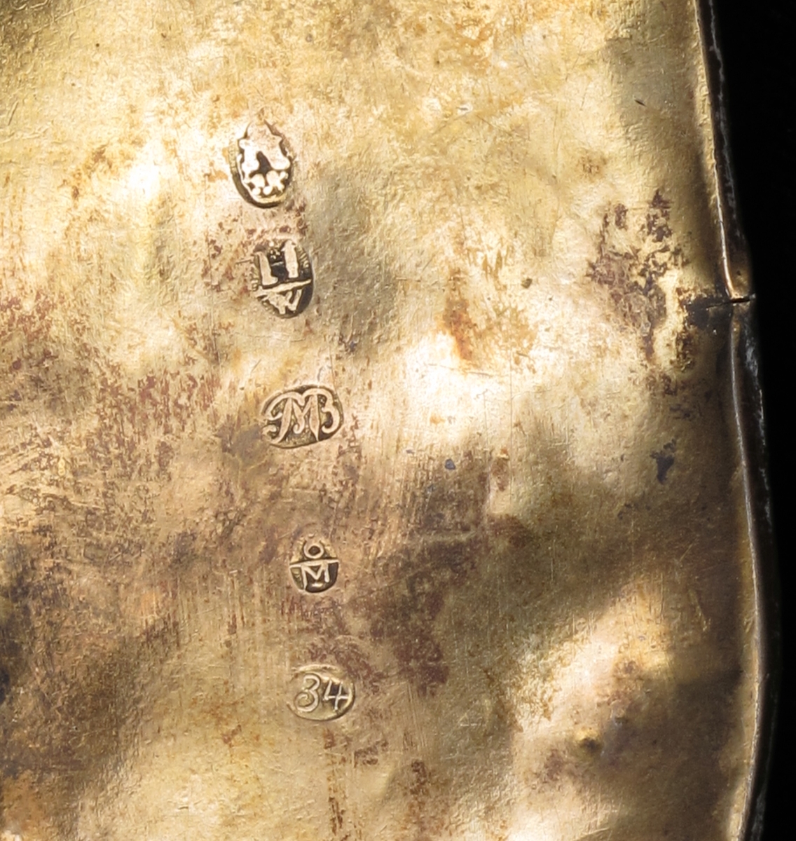 Belte med geheng. Av rød plysj, kantet med smal sort silkebord med små blomsterranker i grønt og fiolett. Beltet besatt med 16 kvadratiske utstansede forgylte sølvplater, hver med to nedhengende løv.  Flatene har rosettornamentikk.  Hektelås av forgylt blikk, de to deler hver med tre glasstener i rødt og grønt.  Ved siden av låsen på den ene side to runde forgylte beslag, utstanset arbeide, med ett løv i sentrum.  Festet til beltet et langt nedhengende geheng av samme materiale, besatt med ni runde utstansede forgylte beslag av forskjellig utførelse og størrelse.  Nederst beslått med avrundet endebeslag, forgylt, med utstanset ornamentikk på oversiden, et ansikt i midten.  Nederst tre løv. Beslaget har gullsmedmerker på baksiden (se Påført tekst) 
Beltet er foret med rødt bomullsstoff med sorte prikker.  Gehenget foret med sort silkebånd, blomsterranke i rosa, og tunget rosa kant.