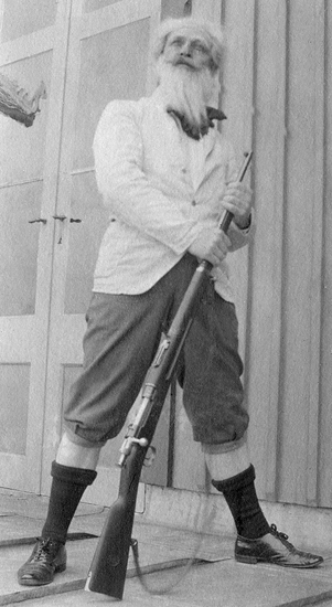 Einar Karl Henrik Nerman (1865-1940), kapten vid Kronobergs regemente, iförd stort vitt lösskägg, klädd i kavaj och knäbyxor m.m. Han står vid en glasdörr och håller i ett gevär av modell 1896.
