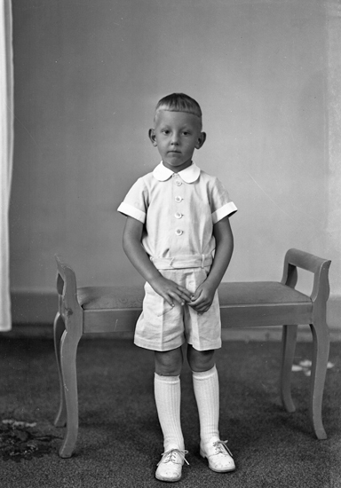 Foto av en pojke i ljus sommarkläder. Han står framför en stoppad bänk. 
Helfigur. Ateljéfoto.