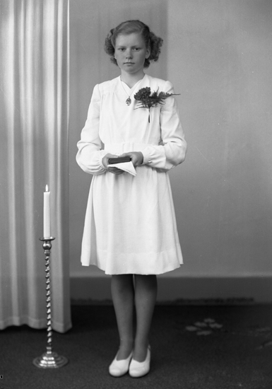 Foto av en ung kvinna i vit konfirmationsklänning. Hon står bredvid en tänd golvljusstake.
Helfigur. Ateljéfoto.