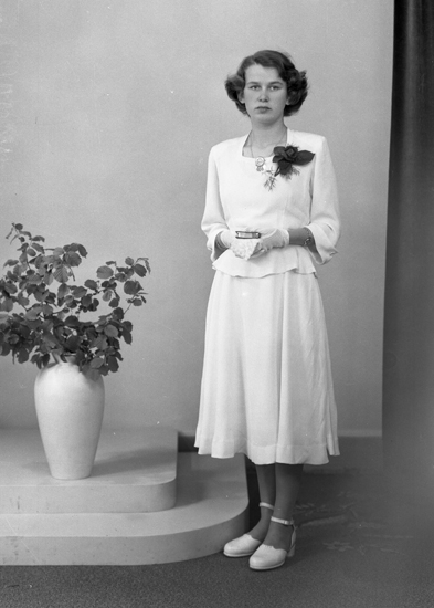 Foto av en ung kvinna i tvådelad vit konfirmationsklänning.
I händerna håller hon en liten psalmbok.
Helfigur. Ateljéfoto.
Konfirmand 1950. Signe Snygg, Hjälmaryd, Blädinge, (1936 - ).
Dotter till Gunnar och Linneá Snygg.