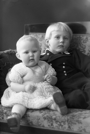 Foto av två små barn, en flicka i stickad klänning och en pojke i kadettuniform. De sitter i en karmstol.
Ateljéfoto.
Karl Gustaf  Petersson (1886-   ), Hjärtenholm, Lekaryd (1930-1935). Gift med Elsa, f. Norinder (1894-   ). Barn: Carl Erik Sölve (1930-   ) och Rut Signe Marianne (1934-   ).
Källa: Församlingsbok, Lekaryd 1924-1941.