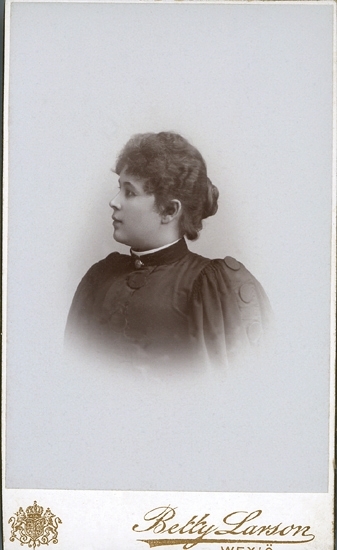 Porträttfoto (bröstbild, profil) av en mörkhårig kvinna i höghalsad, mörk klänning med stora knappar. 
Vid halsen skymtar en brosch.
Självporträtt av fotografen, Betty Larson (1864-1939).