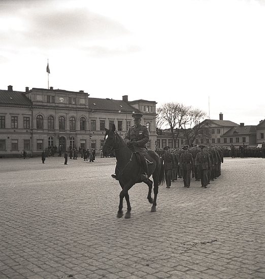 En Krigsmans erinran, 1942.
Regementet marscherar iväg längs Stortorget, framför Residenset. 
I förgrunden syns en officer till häst. I bakgrunden syns Stadshotellet och där skymtar också en del av husen längs Kungsgatan.