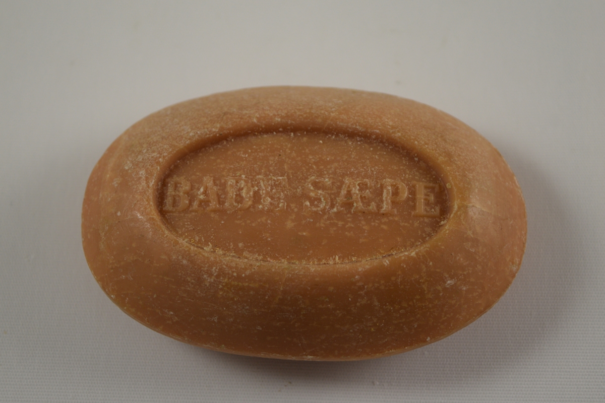 Oval såpe med tekst i relieff i ovale nedsenkede felter på såpens over og underside. Produsert på Saanums Sepefabrikk i Mandal.