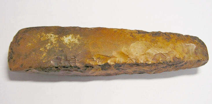Tjocknackig yxa i flinta med gulbrun patina. Slipad egg och delvis slipad kropp. Tydliga repor/slipmärken på yxans sidor. Funnen 1865.