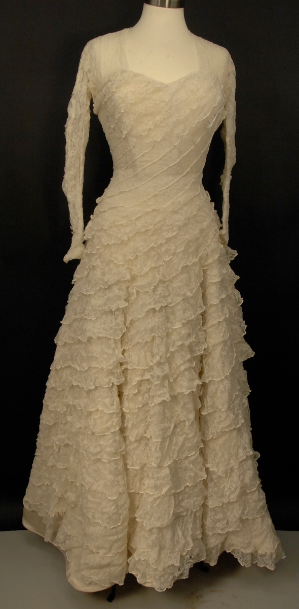 Helklipt, figursydd. Ermer og bærestykket klipt i ett. Rund hals bak med 22,5 cm lang splitt og 14 knapper med sydde trådhemper. Foran er halsutringingen rett nedover 18,5 cm fra skulderbredden ved halsen og går ned under bryststykket på kjolen. Fra bærestykket (som er foret med bobinett) er kjolen foret med tykk, blank silke. Utenpå er påsydd bobinettkapper som går på skrå fra venstre og ned mot høgre. Størsteparten av overdelen og hele skjørtet er blonden sydd på bobinettstoff uten mønster. Underskjørtet er da løst og har ei snor innsydd i folden nede for at kjolen skal føre seg penere. Glidelås i siden, hekter og sydde hemper på overdelen. Erme: Skinkeerme med 12,5 cm lang splitt ved håndleddet og 7 knapper, sydde knappehemper. 6 små innsydde legg på underarmen for bue av ermet. Skjørtet er utsvingt og har ein vidde nede på ca. 668 cm. Vidden på underskjørtet 470 cm. Kjolen er 19 cm lengre bak enn foran.