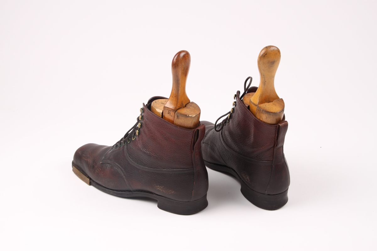 Et par beksømstøvler i lær med maljer i metall samt messingbeslag på sålen. Støvlene har lærlisser med sølvbeslag og en hempe av skinn bak på hver sko. Trelester inne i skoene.