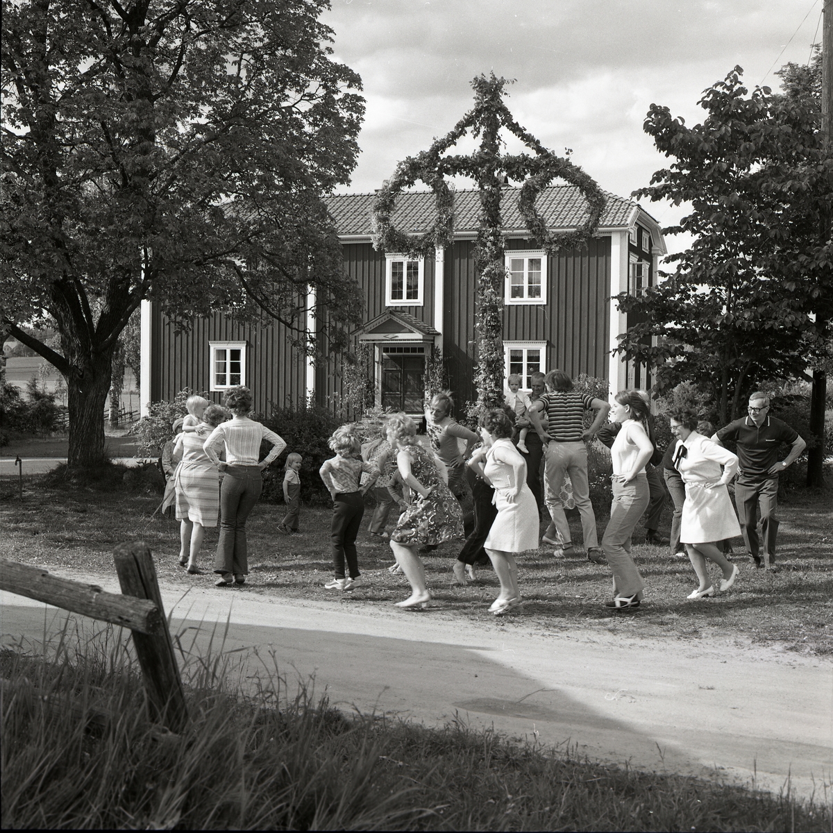 Dom äro verkligen lustiga att se, de som dansar små grodarna runt midsommarstången i Glössbo 1972. Framför bostadshuset och trädgården har midsommarstången rests och ramas in av omkringliggande lövträd med full sommarskrud.