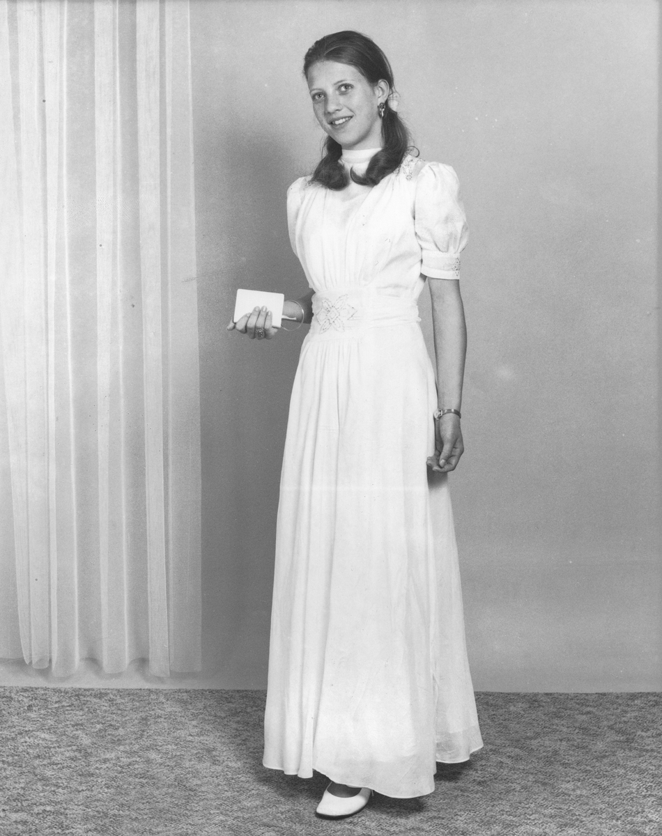 Studioportrett av konfirmant i hvit kjole og med salmebok i handa.
