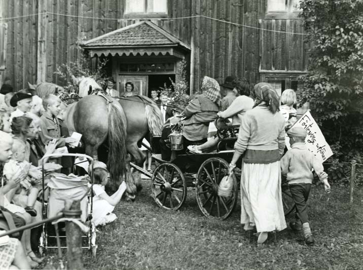 En vagn dragen av två hästar mot en välbesökt hembygdsgård.