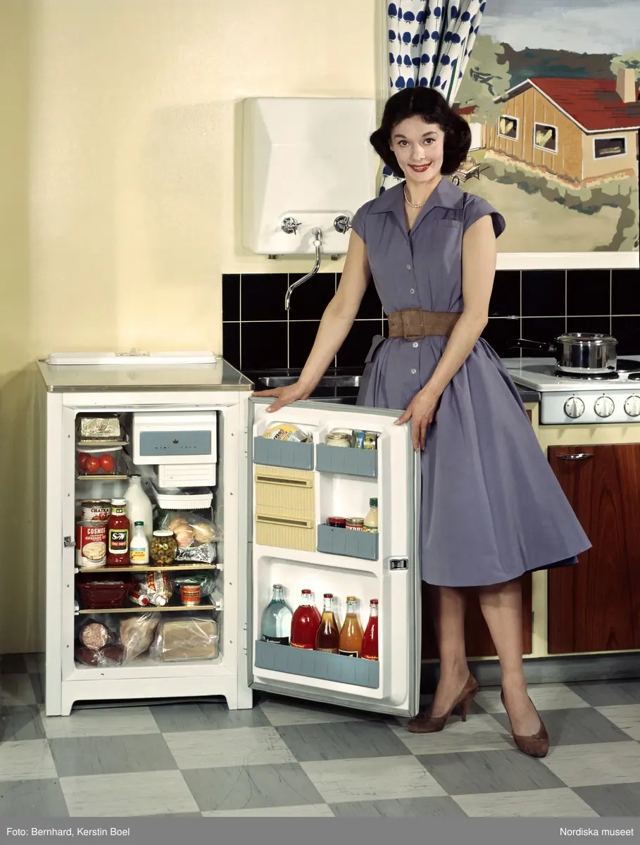 Kvinna i köksmiljö. Kylskåp med öppen dörr, fyllt med matvaror.