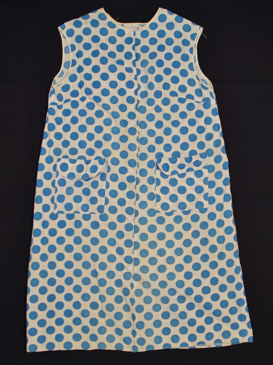 Ermlaus kjole med opning midt framme. Lukking med knappar. to lommer. Kvitt stoff med store, blå prikkar.