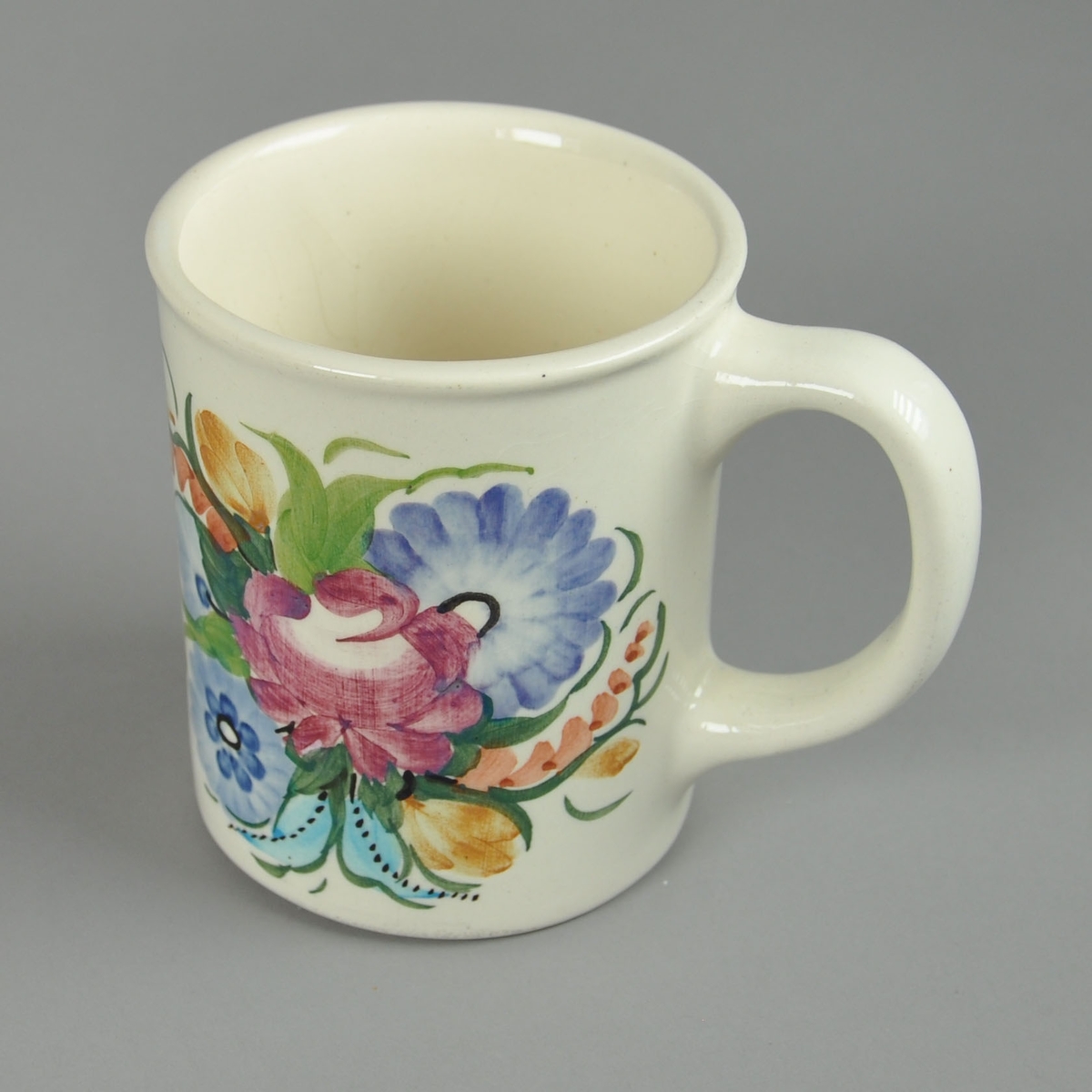 Kopp av glassert keramikk, med malt dekor. Motivet på dekoret er blomsterkrans på to sider av koppen. Koppen har hank.