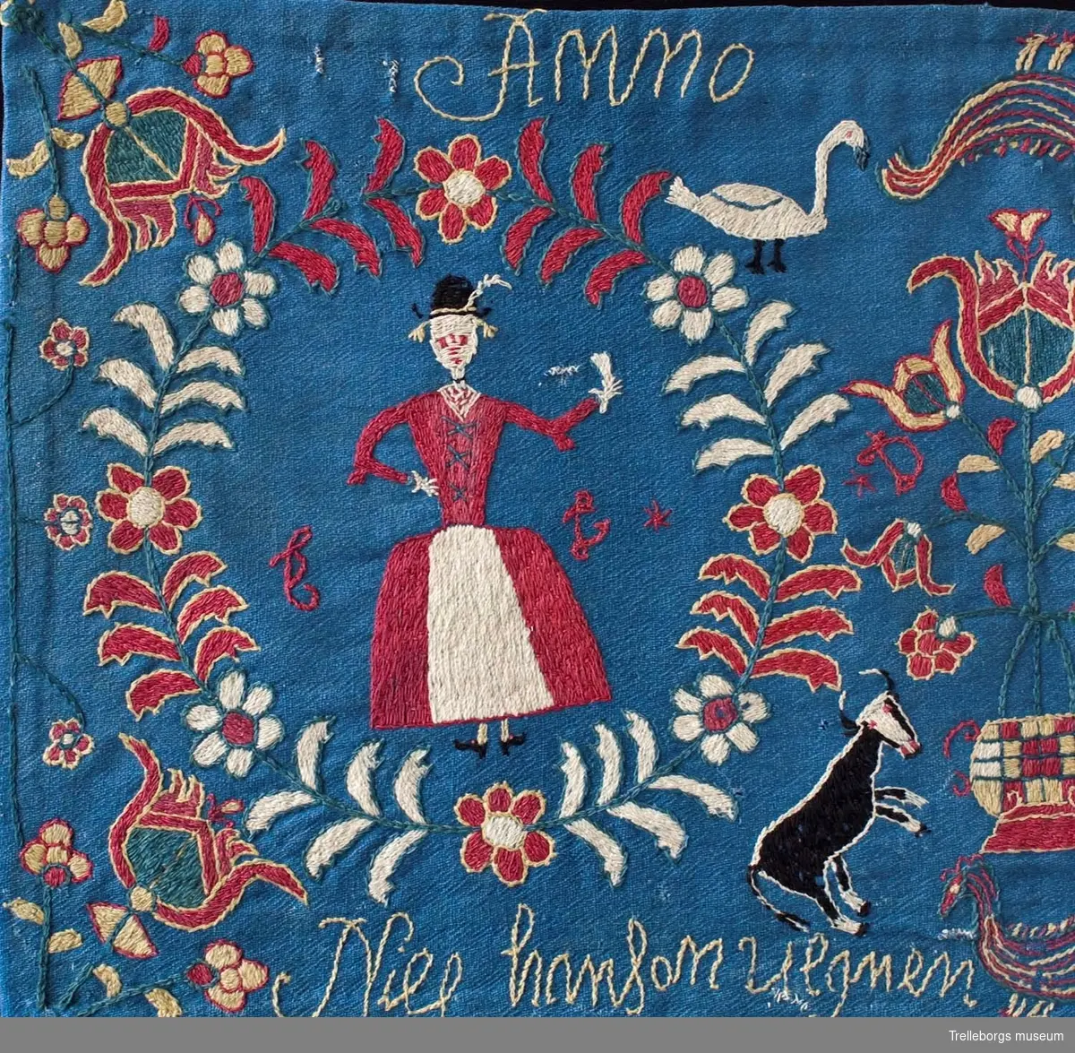 På en mellanblå botten är två kransar broderade, i den ena kransen står en kvinna och i den andra en man. I dynans hörn är blommor broderade, samma blomma kommer igen i dynans mitt tillsammans med en tupp, två gäss och två kor. Broderiets färger är rött, vitt, gult, grönt och svart. Dynan är märkt Anno 1780 Nils Hansson Ulgren Kierstin Andersdotter.