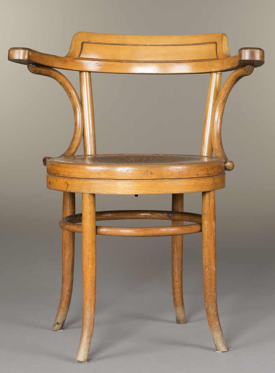 Kontorstol i lyst treverk, med armlener. Stolen har elementer av bøyd trearbeid. Et rundt svingsete. Ryggstykket har enkel linjedekor som følger formen. Setet har et utskjært mønster i form av bladverk.