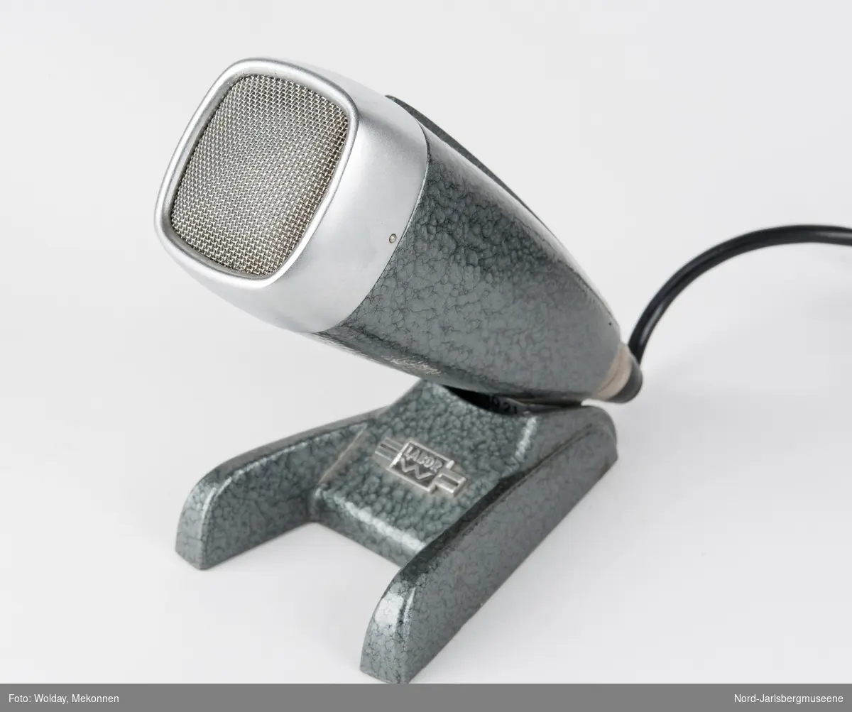En kantet konisk mikrofon på en støttefot. Hodet er blankt metall, resten er i hamret, eloksert aluminium. Påskrudd svart koblingsledning for høyttaler.