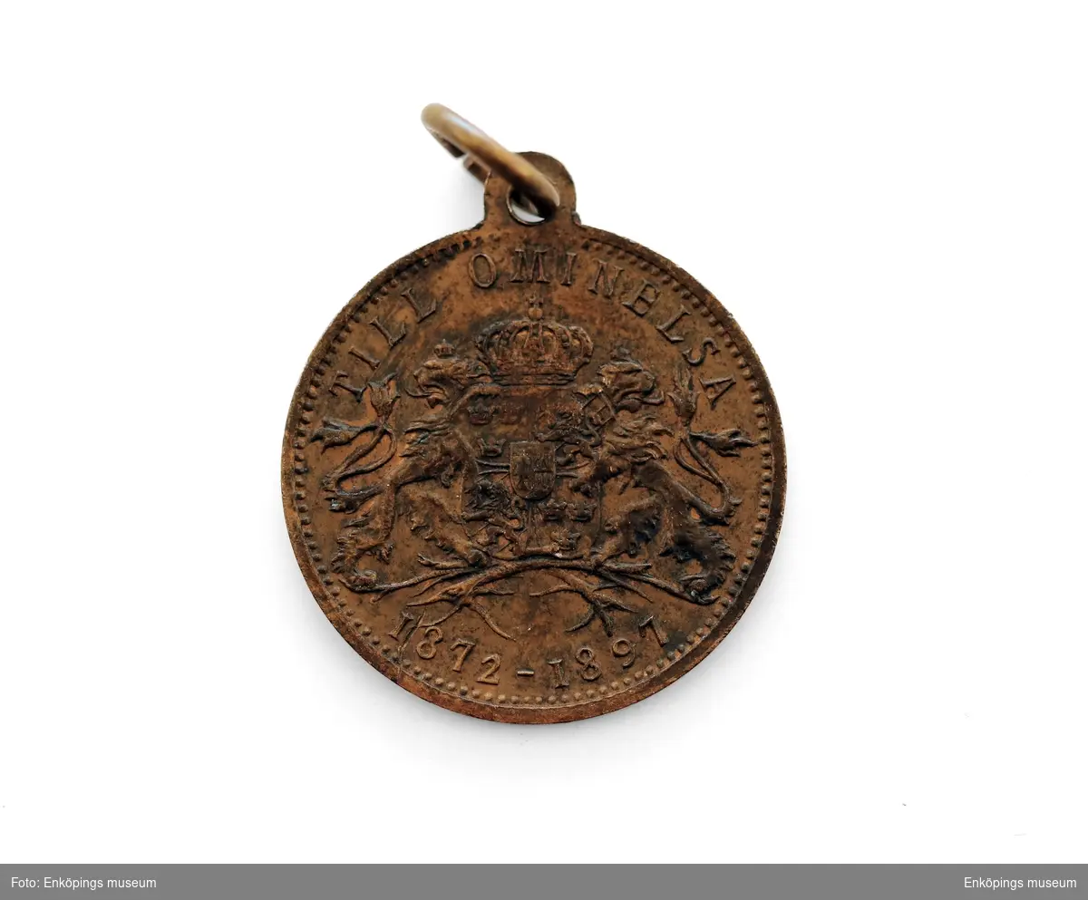 Rund medalj av koppar med hänge. Profilbild av kung med texten: " OSCAR II KONUNG SVERIGE O NORGE". På baksidan ett krönt vapen med texten: " TILL OMINELSE" " 1872- 1897".  Del av en samling med 7 medaljer sammanbundna på en ring till en knippa.