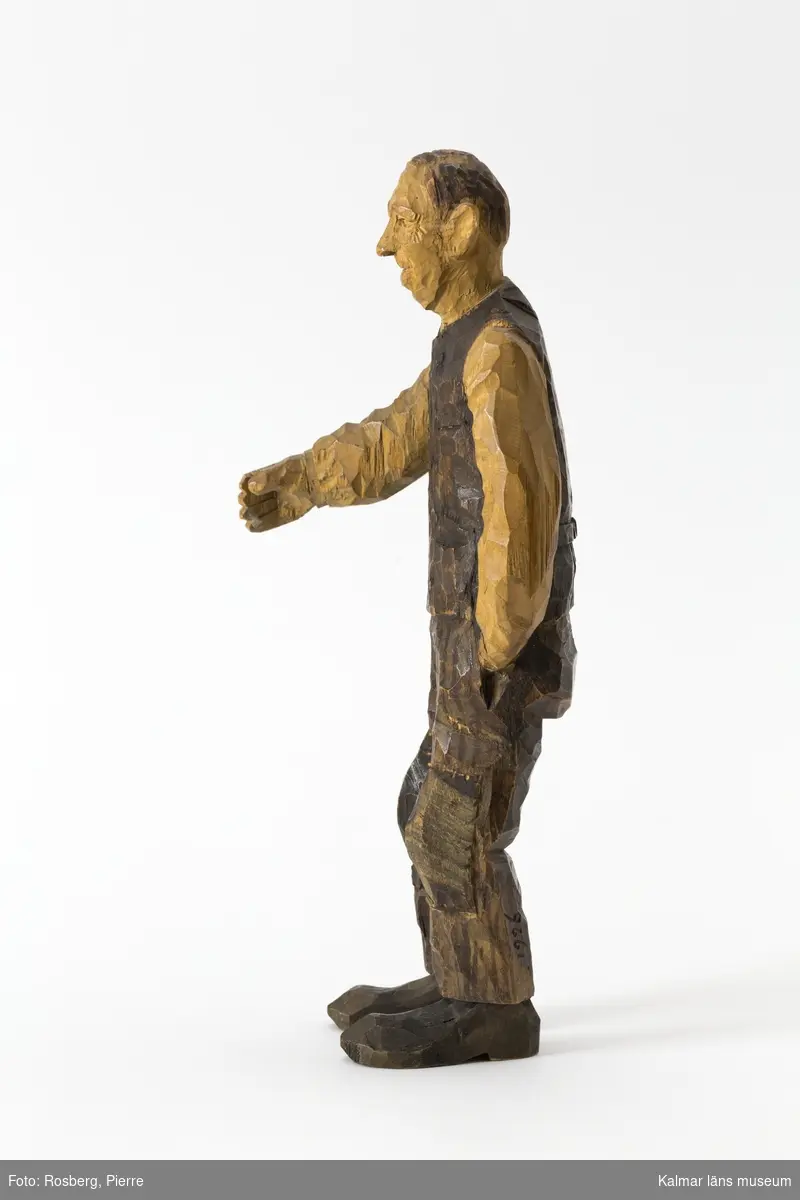 KLM 45932:1 Skulptur, av trä, målad. Figur i form av en stående man. Han har vänster hand i byxfickan och den högra armen sträcker han fram, möjligen har han ursprungligen haft något föremål i den framsträckta handen, barhuvad, klädd i skjorta väst och byxor, stora lappar på knäna, och skor. Signerad på ryggen, H.C., på vänster byxben inskuret, 1926. Målad i bruna nyanser. Tillverkad av Helge Rugland, f. Carlsson.