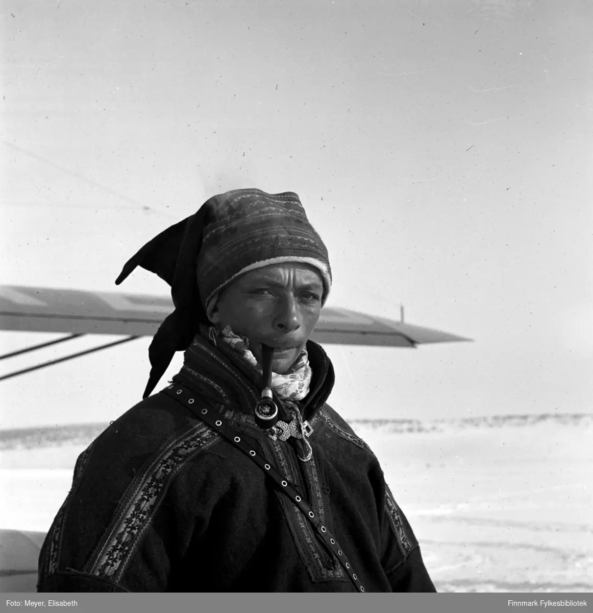 Johan Mathisen Hætta med pipe, fotografert foran flyet som landet på Kautokeinoelva.