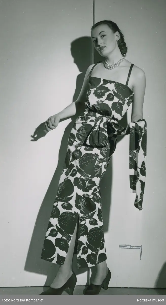 Modell i stormönstrad klänning med slits och midjeband, pärlhalsband och pumps, från Edward Molyneux.