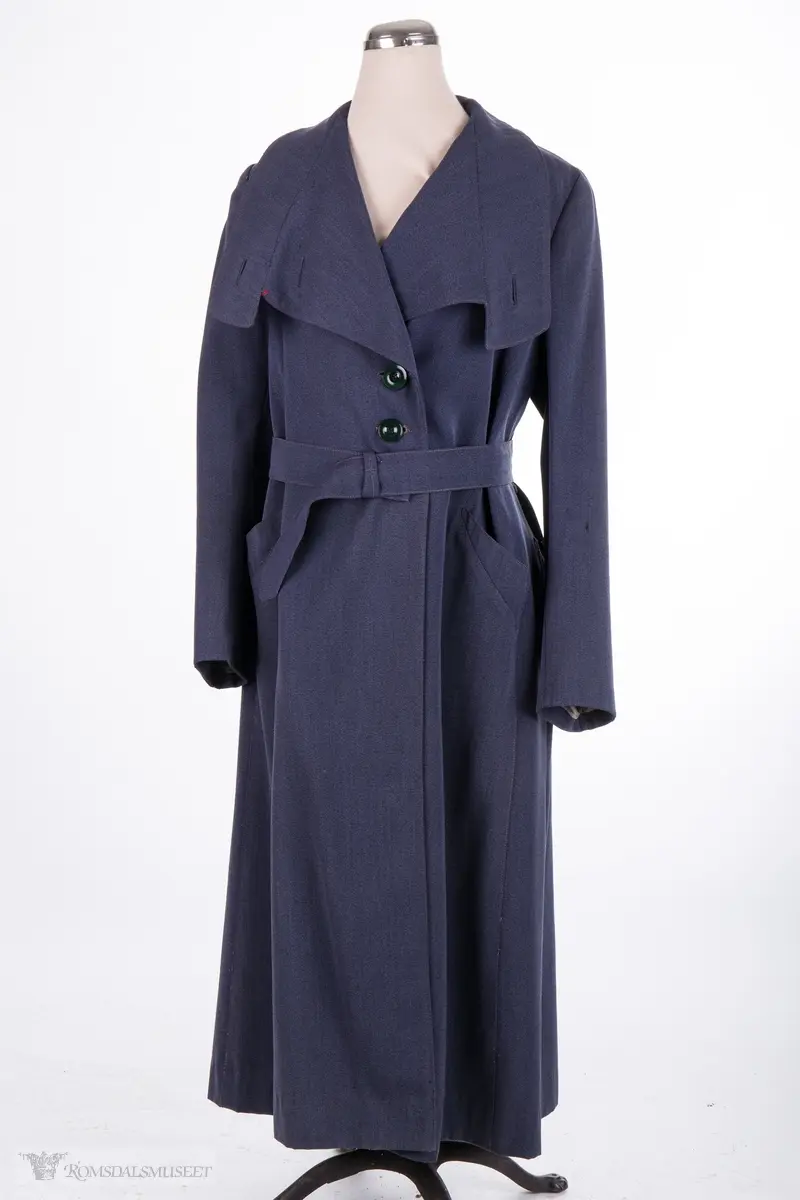 Kappe med store slag. Lukkes over brystet med to store knapper. To skrå stikklommer, lange ermer. Belte med spenne hører til.