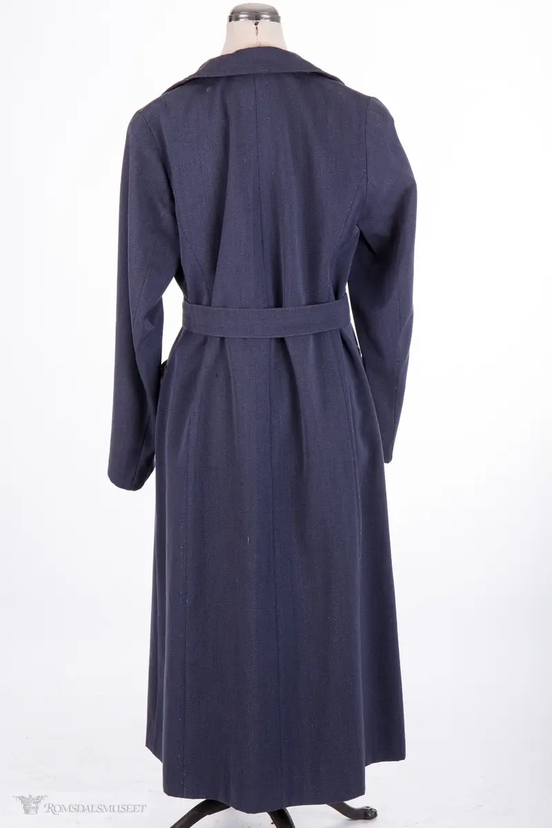 Kappe med store slag. Lukkes over brystet med to store knapper. To skrå stikklommer, lange ermer. Belte med spenne hører til.