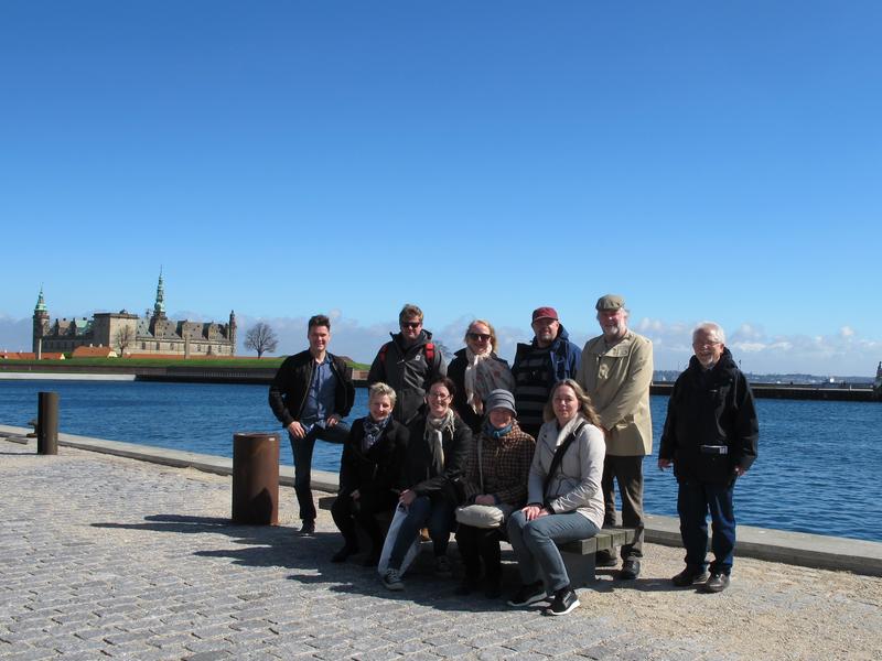 Ti mennesker på og rundt benk i forgrunnen, Kronborg slott ved Øresund, Danmark i bakgrunnen.